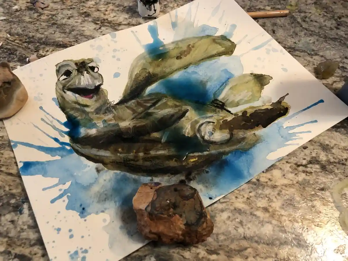 A watercolor painting of a frog with a wide, smiling mouth and large eyes, lying on its back in a pool of blue water…