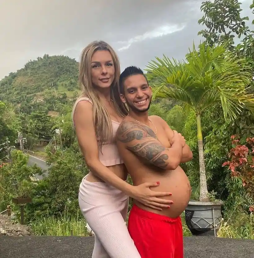 A man with a visibly pregnant belly, wearing red shorts and a tattooed arm, stands smiling with his arms crossed while a…