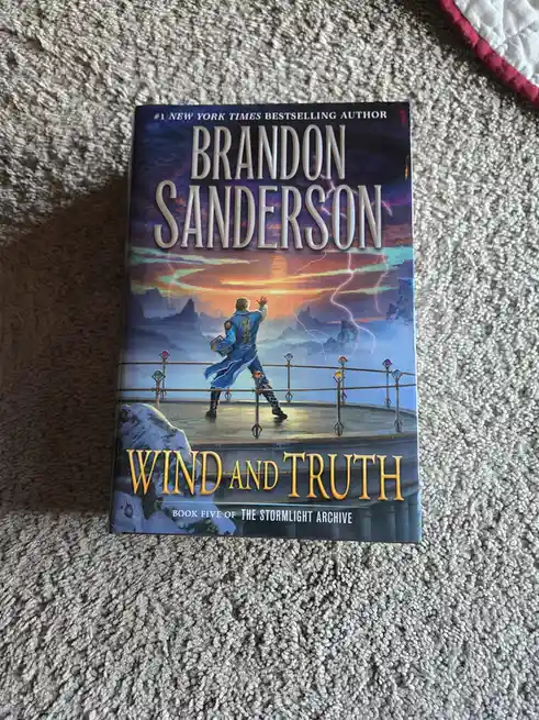 A physical copy of Brandon Sanderson's book 'Wind and Truth,' book five of 'The Stormlight Archive,' rests on a textured…