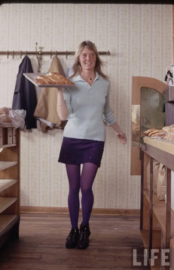 A woman in a light blue sweater and purple skirt and tights stands in a bakery holding a tray of bread. She smiles at the…