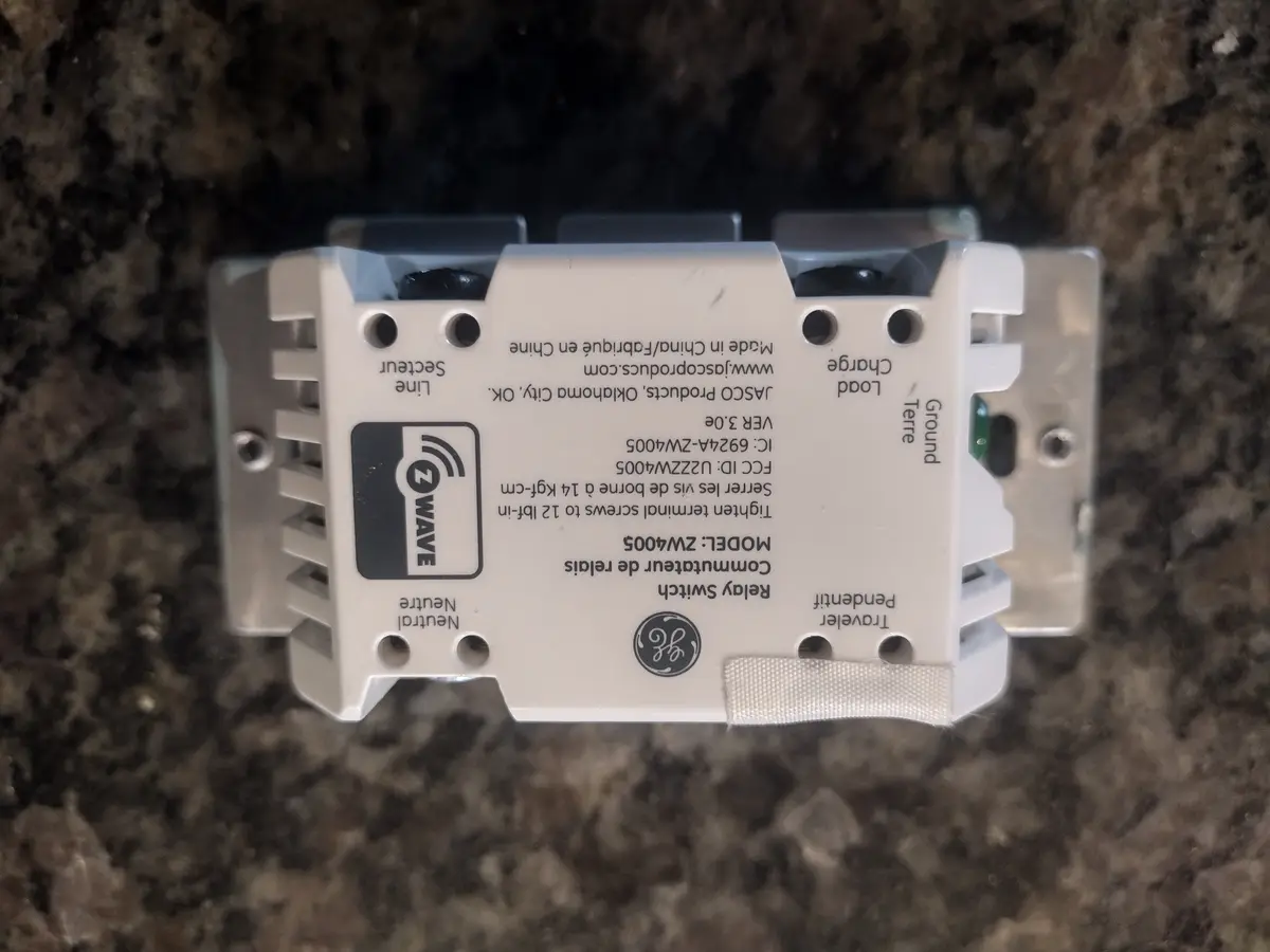 A close-up photo of a white GE Z-Wave relay switch, model ZW4005, lying on a dark speckled countertop. The device has…
