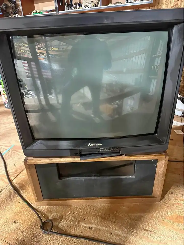 A black Mitsubishi CRT television sits on a wooden stand in a cluttered room, its screen dark and reflecting a blurry…