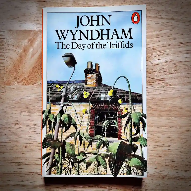 A physical copy of John Wyndham's novel 'The Day of the Triffids' is shown lying flat on a wooden surface. The book cover…