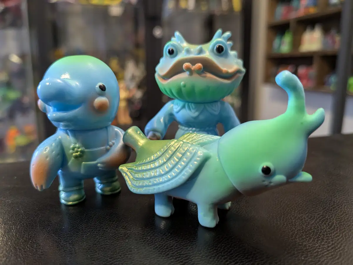 Three blue-green ceramic figurines, resembling stylized sea creatures or mythical beings, are arranged on a dark textured…