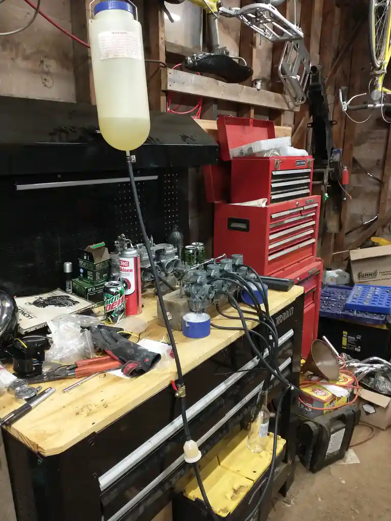A cluttered garage workshop features a wooden workbench covered with tools, carburetors, a can of Mountain Dew, and a red…