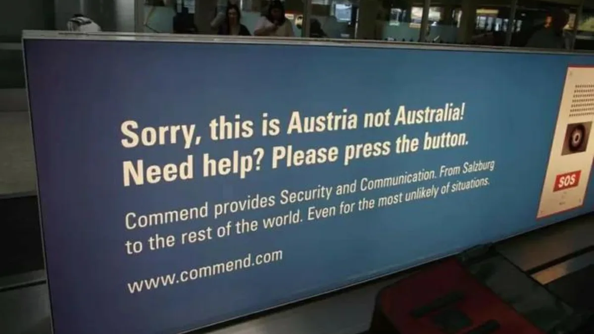 A blue airport sign with white text humorously corrects a common traveler mistake by stating 'Sorry, this is Austria not…