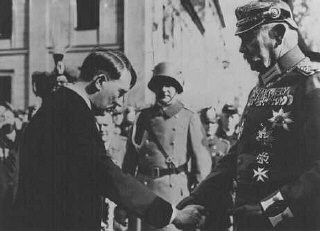 A black-and-white historical photograph shows Adolf Hitler shaking hands with a high-ranking German military officer adorned…