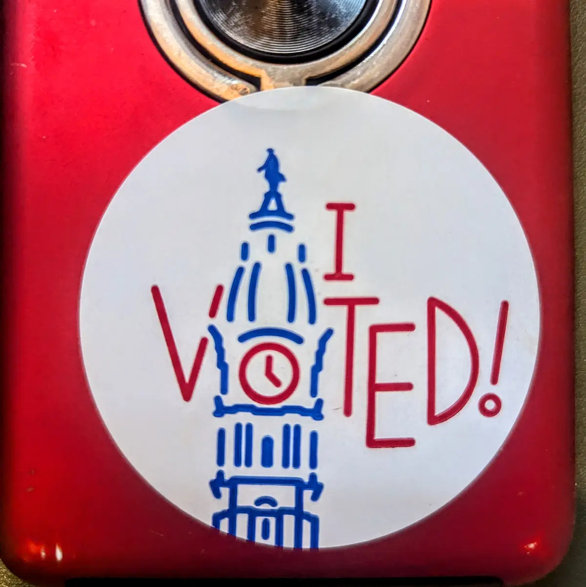 A close-up of a red device, likely a phone or camera, with a white circular sticker on it. The sticker features a blue…