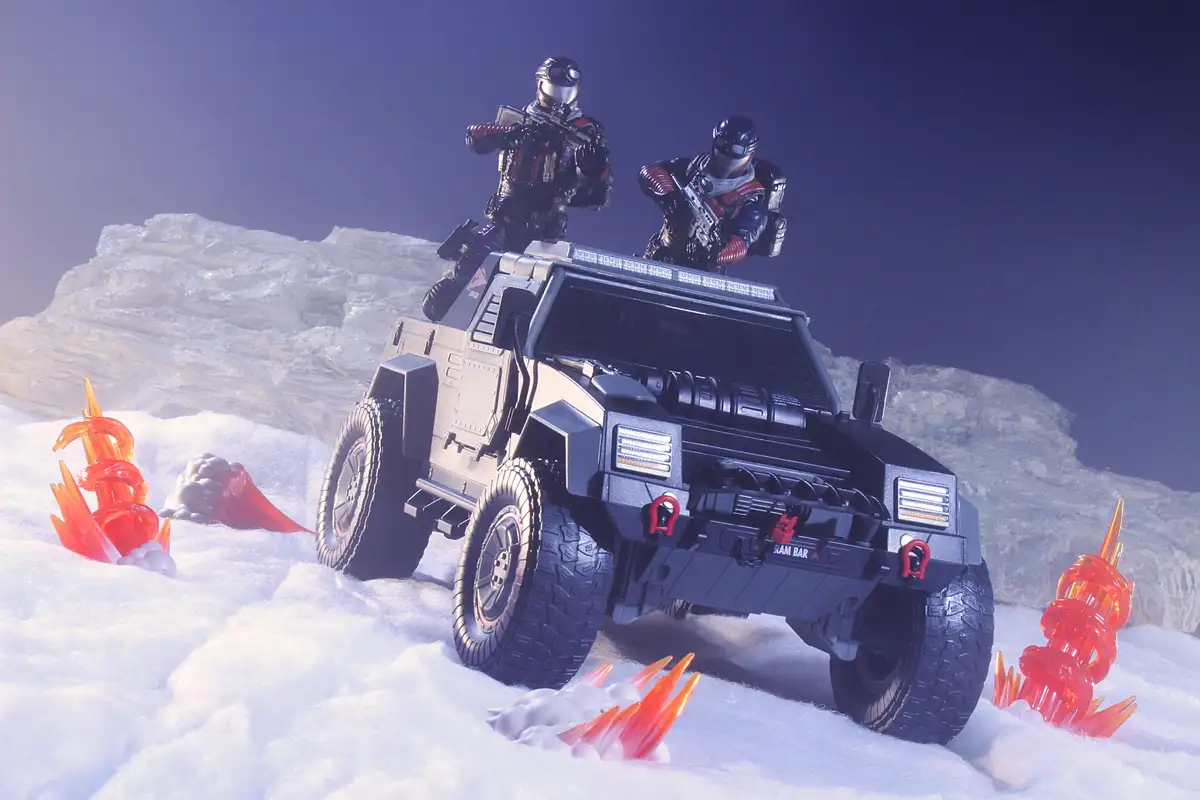 A toy military vehicle with two armed toy soldiers on its roof is shown in a snowy landscape with plastic orange flame-like…