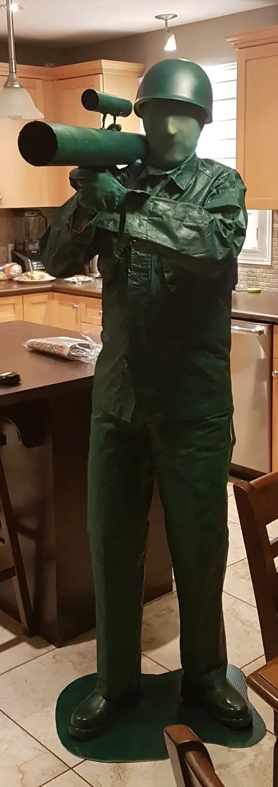 A full-body green toy soldier figure, complete with helmet and a large green toy rocket launcher, stands in a kitchen. The…