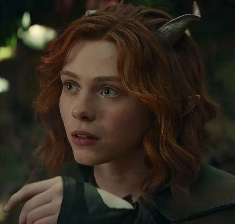 A close-up of a woman with red hair, small horns, and pointed ears, looking surprised or confused. She wears a dark green…