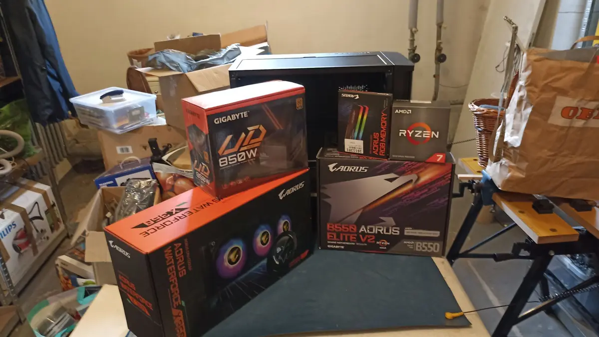 A cluttered garage or workshop scene shows several unopened computer component boxes from brands like Gigabyte, Aorus, and…