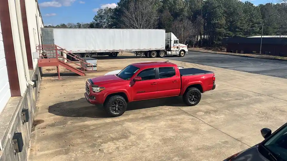 A red Toyota Tacoma pickup truck is parked in front of a warehouse with a large white semi-truck trailer visible in the…