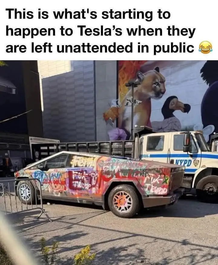 A heavily graffitied Tesla Cybertruck is parked on a city street next to an NYPD truck, with visible tags like 'FEM' and…