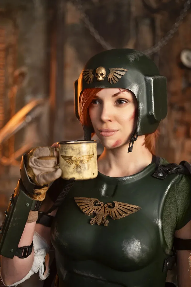 A woman in a green armored costume with a skull-and-wings emblem on her helmet and chestplate holds a matching coffee mug…