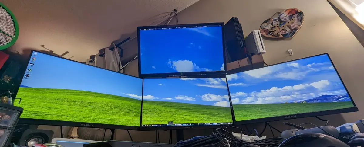 Four computer monitors arranged in a curved cluster display the iconic Windows XP 'Bliss' wallpaper of a green field under a…