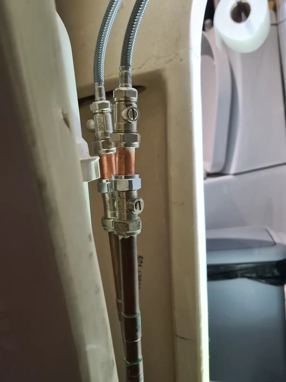 A close-up photo shows plumbing pipes, including copper and braided metal hoses, connected to a toilet tank. A roll of…