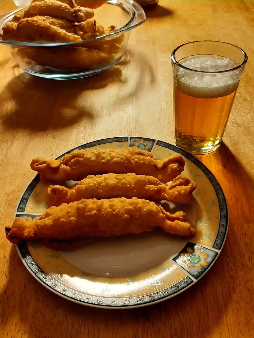 A wooden table holds a plate with three golden-brown, crispy fried chicken tenders and a glass of beer with a frothy head…