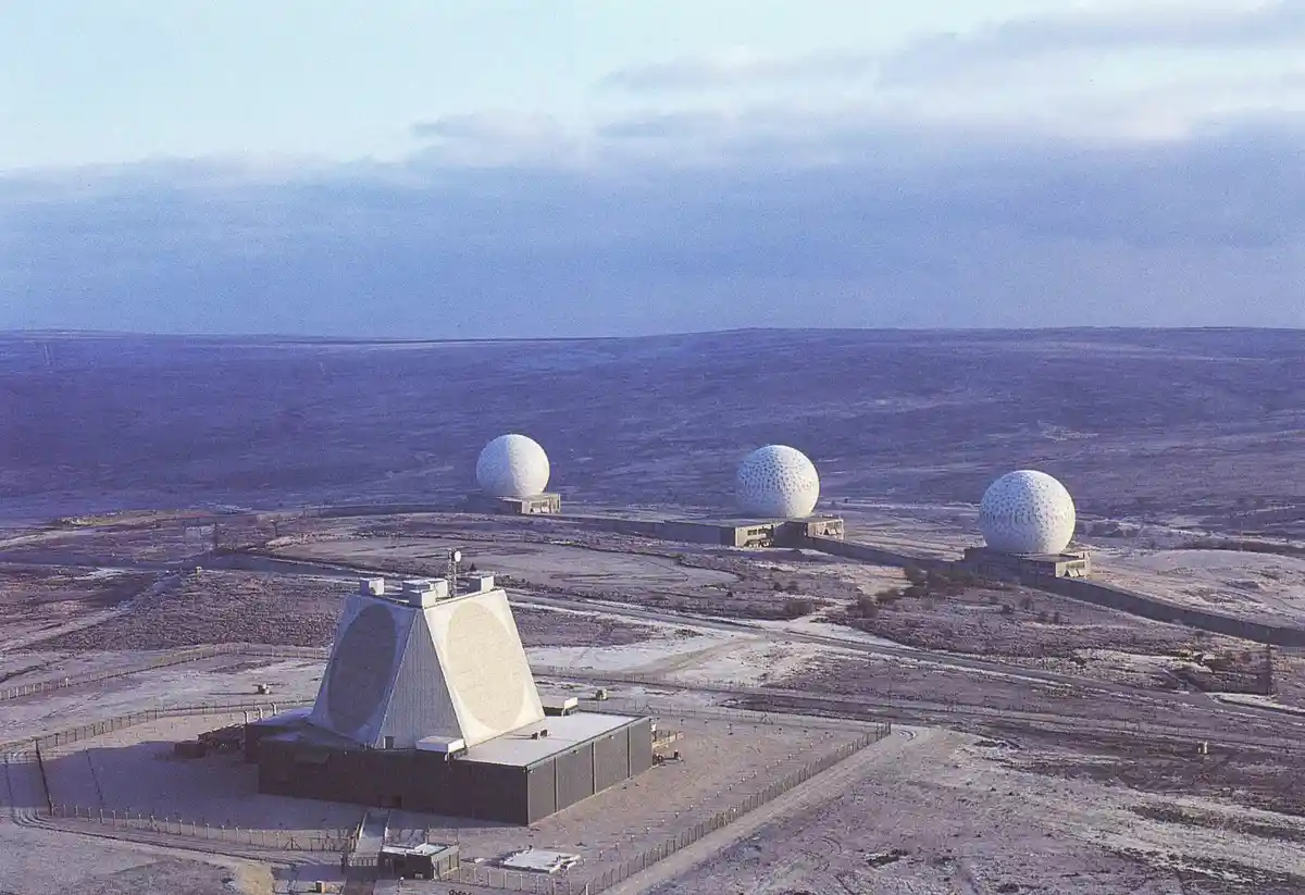 An aerial view of a desert radar installation featuring a large, angular main radar dish in the foreground and three…