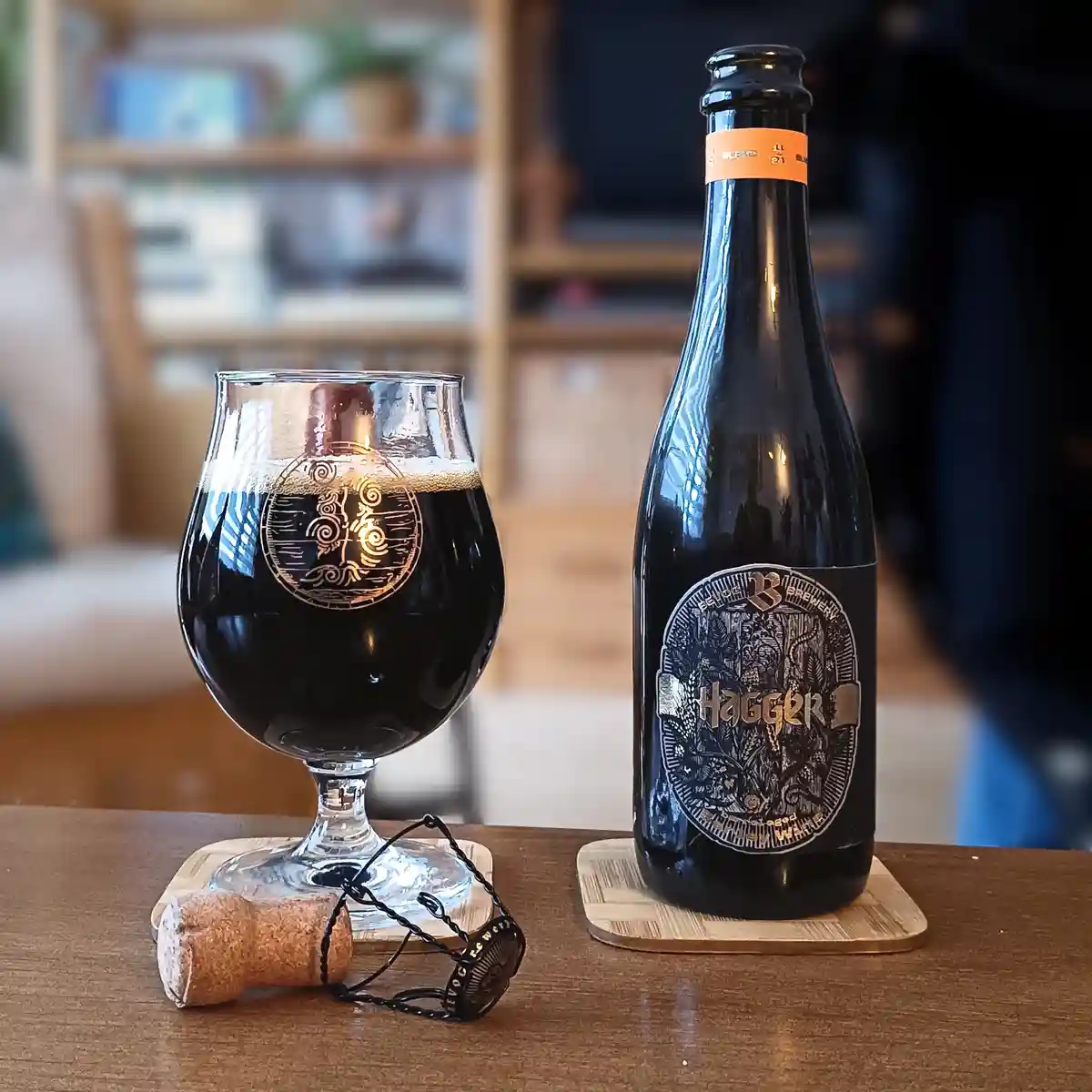 A dark beer bottle labeled 'Hagger' from Bevoc Brewery sits next to a filled snifter glass on wooden coasters, with a cork…