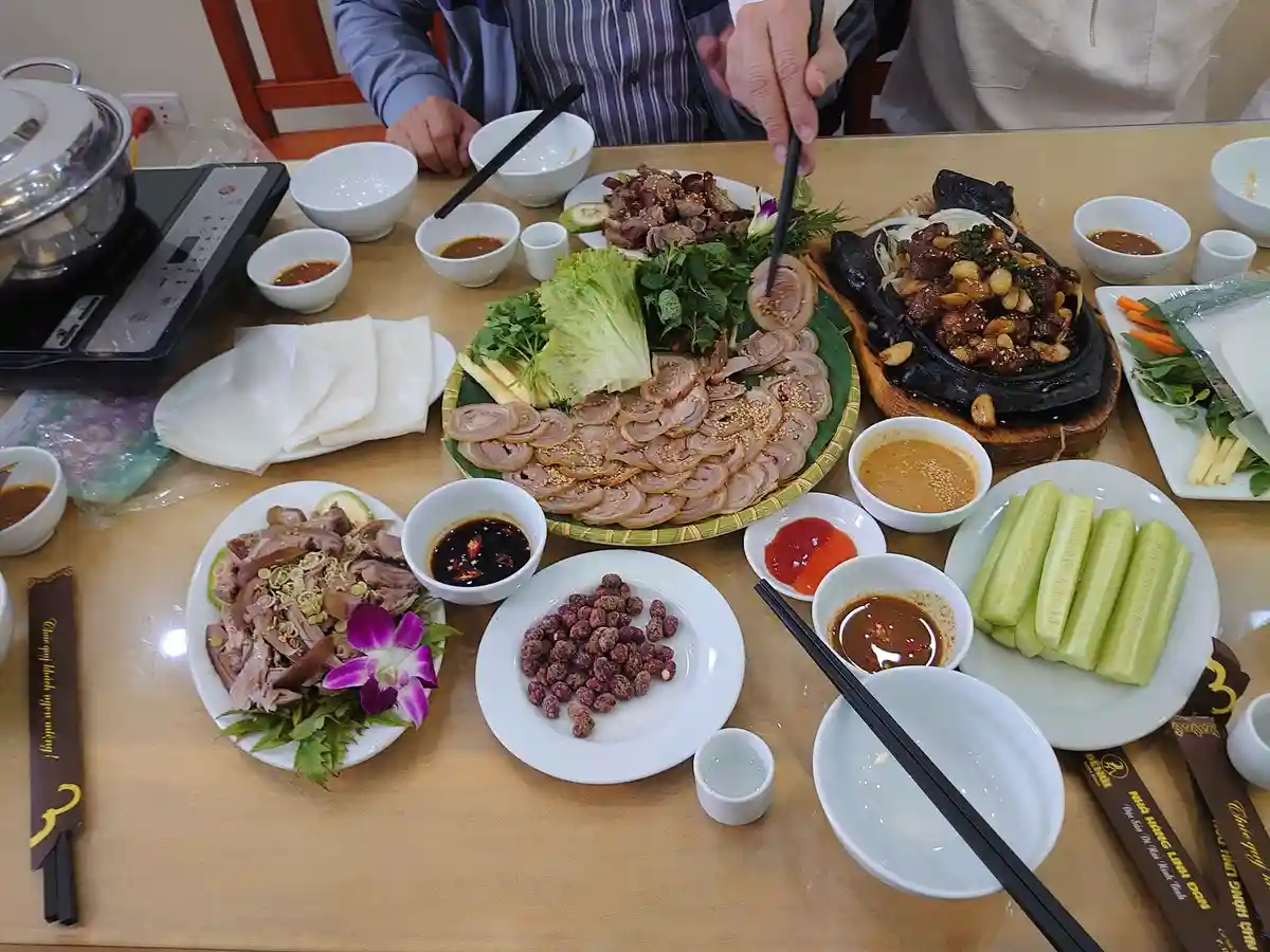 A table laden with an abundant Vietnamese feast featuring sliced meats, fresh herbs, cucumbers, sauces, and a sizzling hot…