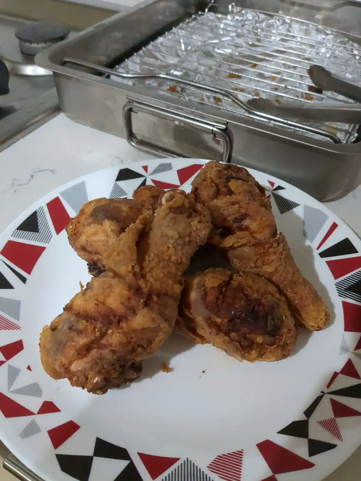 A plate of golden-brown fried chicken pieces sits on a white plate with a red, black, and gray geometric pattern. In the…