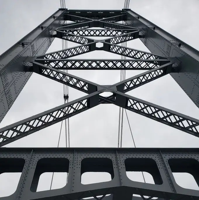 A low-angle black-and-white photograph of a steel truss bridge's intricate lattice structure against a cloudy sky. The image…