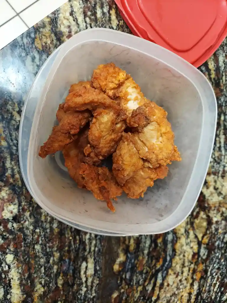 A clear plastic container filled with golden-brown fried chicken pieces sits on a speckled granite countertop, with a red…