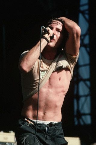 A shirtless man with a ripped shirt, holding a microphone to his mouth while raising his other hand to his head, appears…