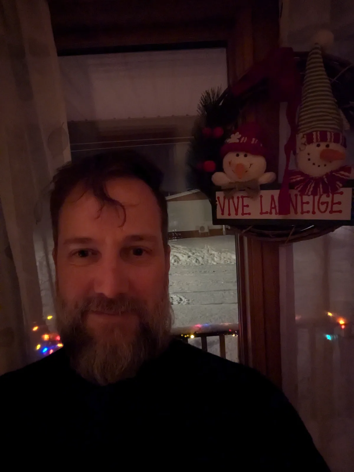A man with a beard takes a selfie indoors near a window showing a snowy scene outside. Above him, a festive wreath with two…
