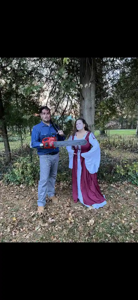 Two people stand outdoors in a wooded area with fallen leaves. The man on the left wears a blue shirt and gray pants, has…