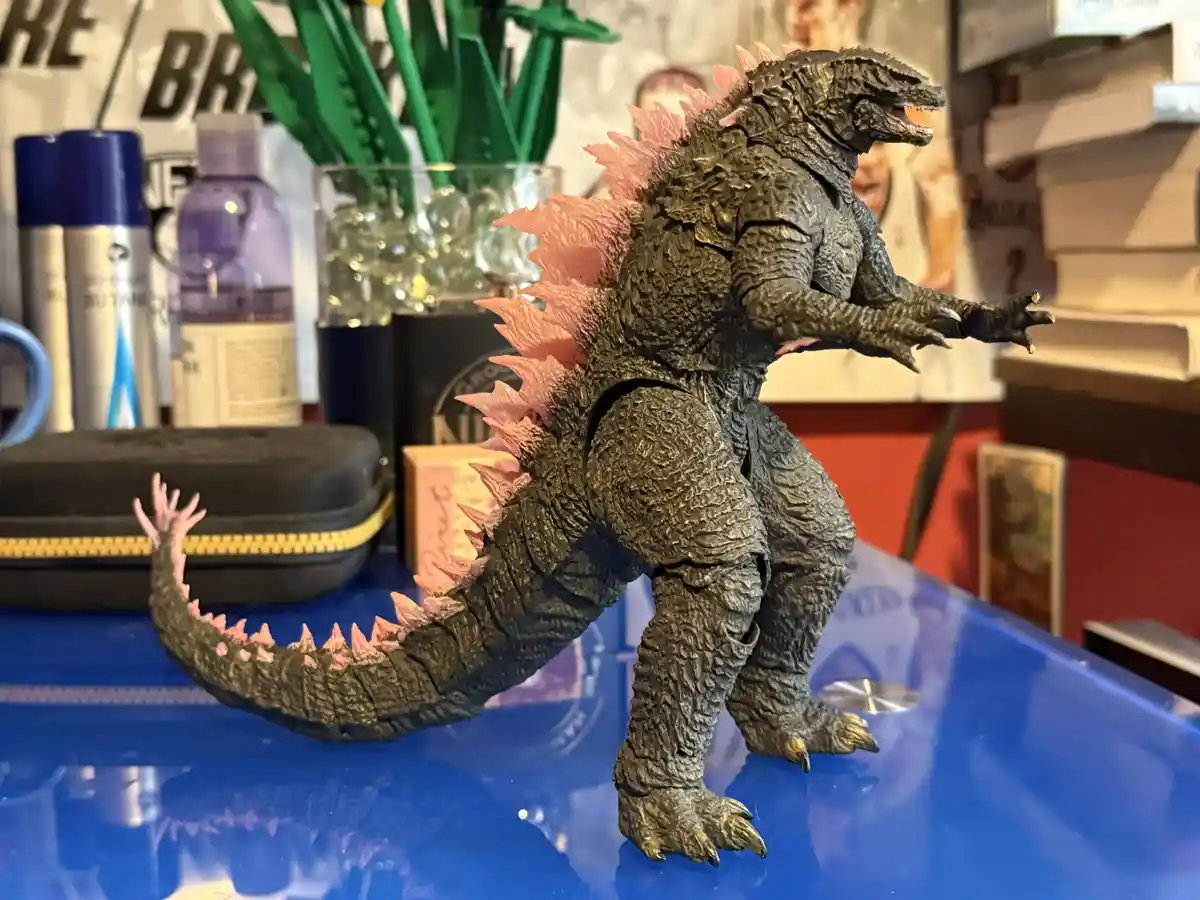 A detailed Godzilla action figure with textured gray skin and pink dorsal fins stands on a blue surface, surrounded by…