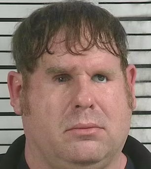 A mugshot-style photo of a man with messy hair and a neutral expression, standing in front of a striped background. The…