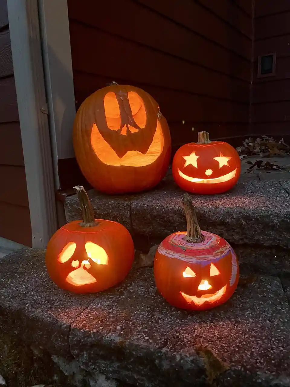 Four carved pumpkins with lit interiors sit on stone steps outside a house, each with a different facial expression. The…