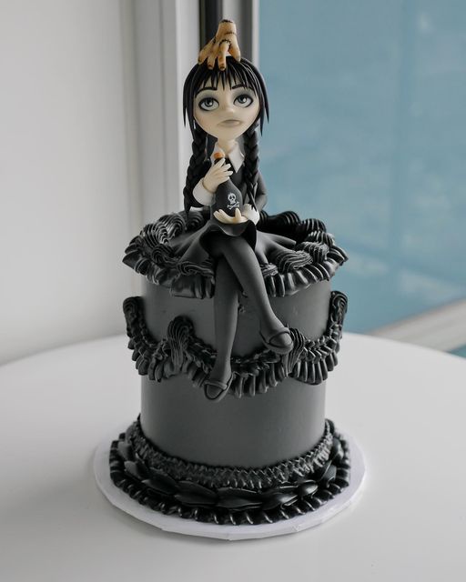 A black-frosted cake features a detailed fondant figurine of Wednesday Addams sitting on top, holding a small skull bottle…