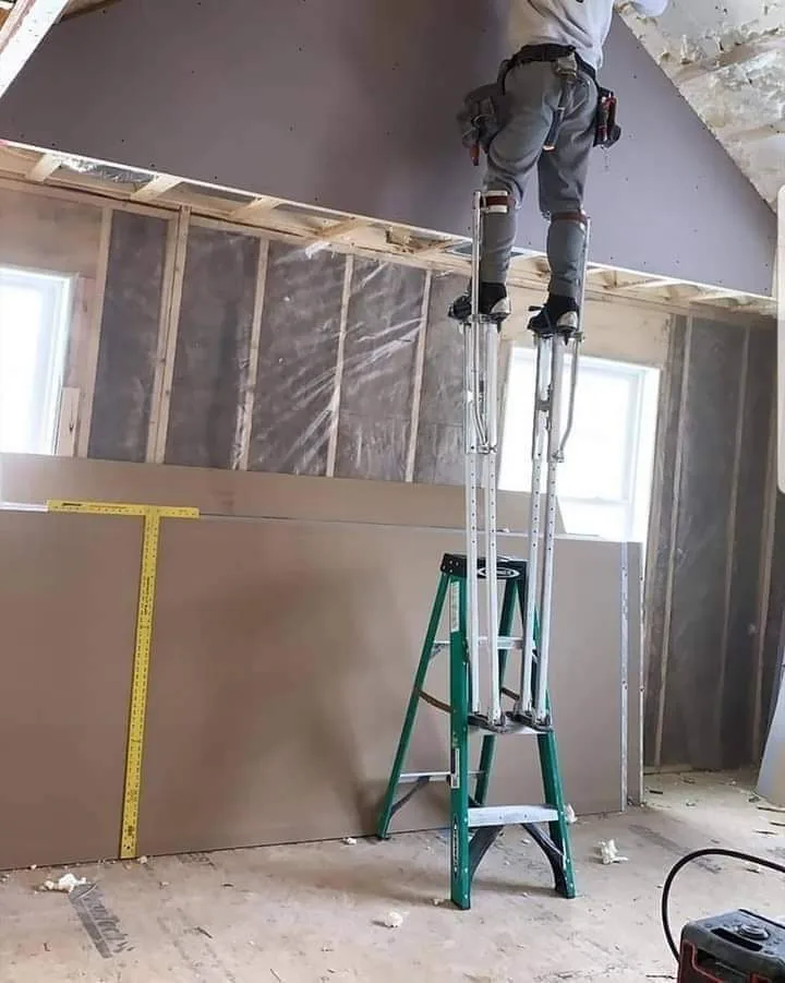 A construction worker stands on a tall, silver telescoping ladder, reaching high to work on a ceiling in an unfinished room…