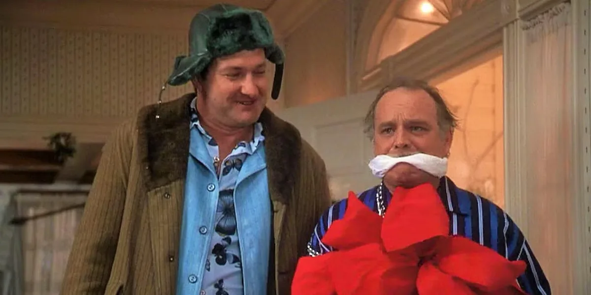 Two men stand indoors; the man on the left wears a green fur hat and brown coat over a blue floral shirt, smiling slightly…