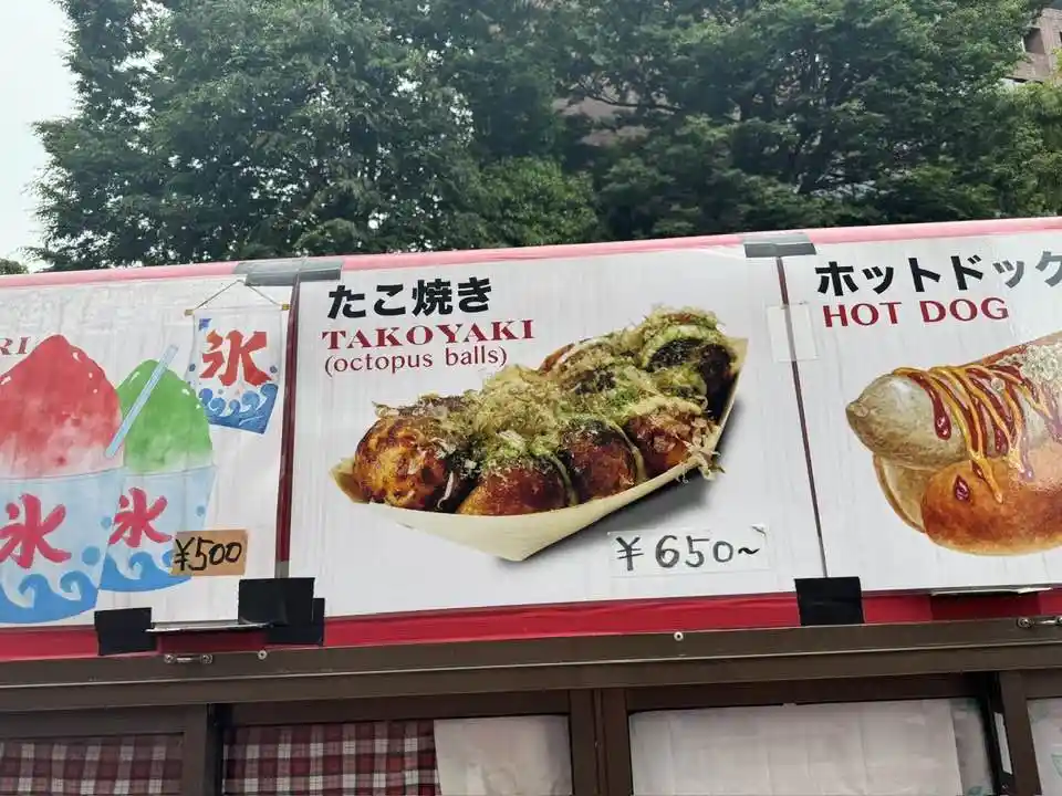 A food stall sign in Japan displays three menu items: shaved ice for ¥500, takoyaki (octopus balls) for ¥650, and a hot dog…