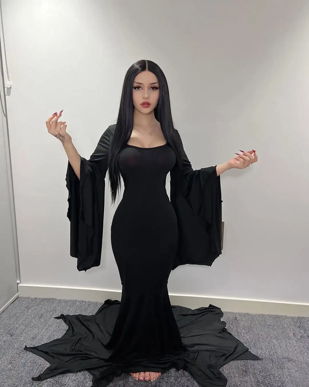 A woman with long black hair and dramatic makeup stands in a form-fitting black dress with wide, flowing sleeves, posing…