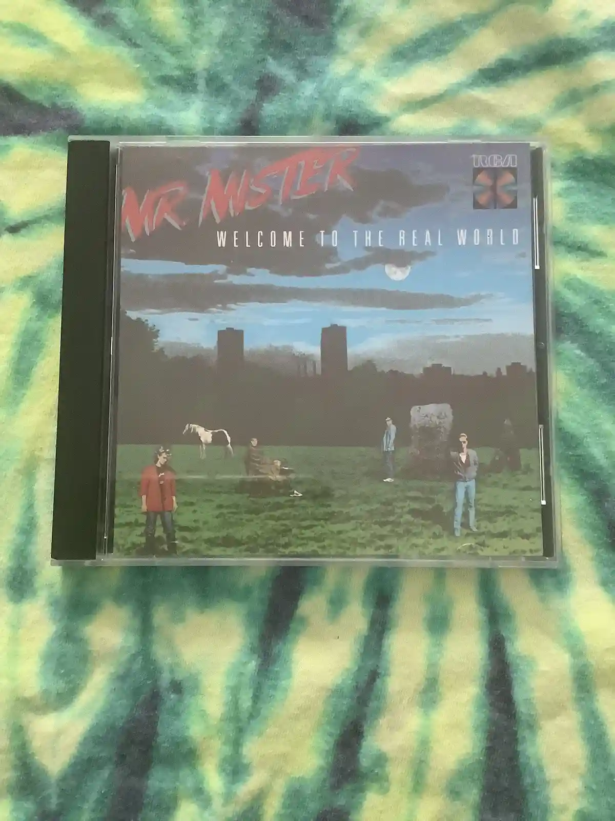 A CD case for the album 'Welcome to the Real World' by Mr. Mister is shown, featuring an illustrated cover with several…