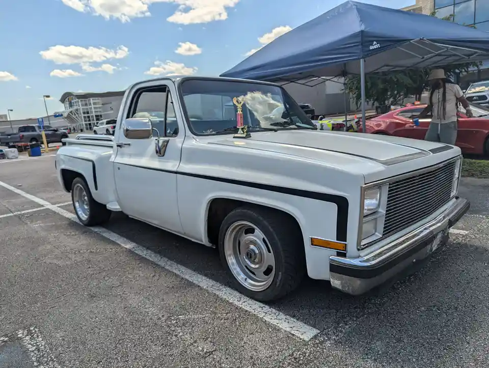 A white classic Chevrolet pickup truck is parked in an outdoor lot under a blue canopy, with a small trophy placed on its…