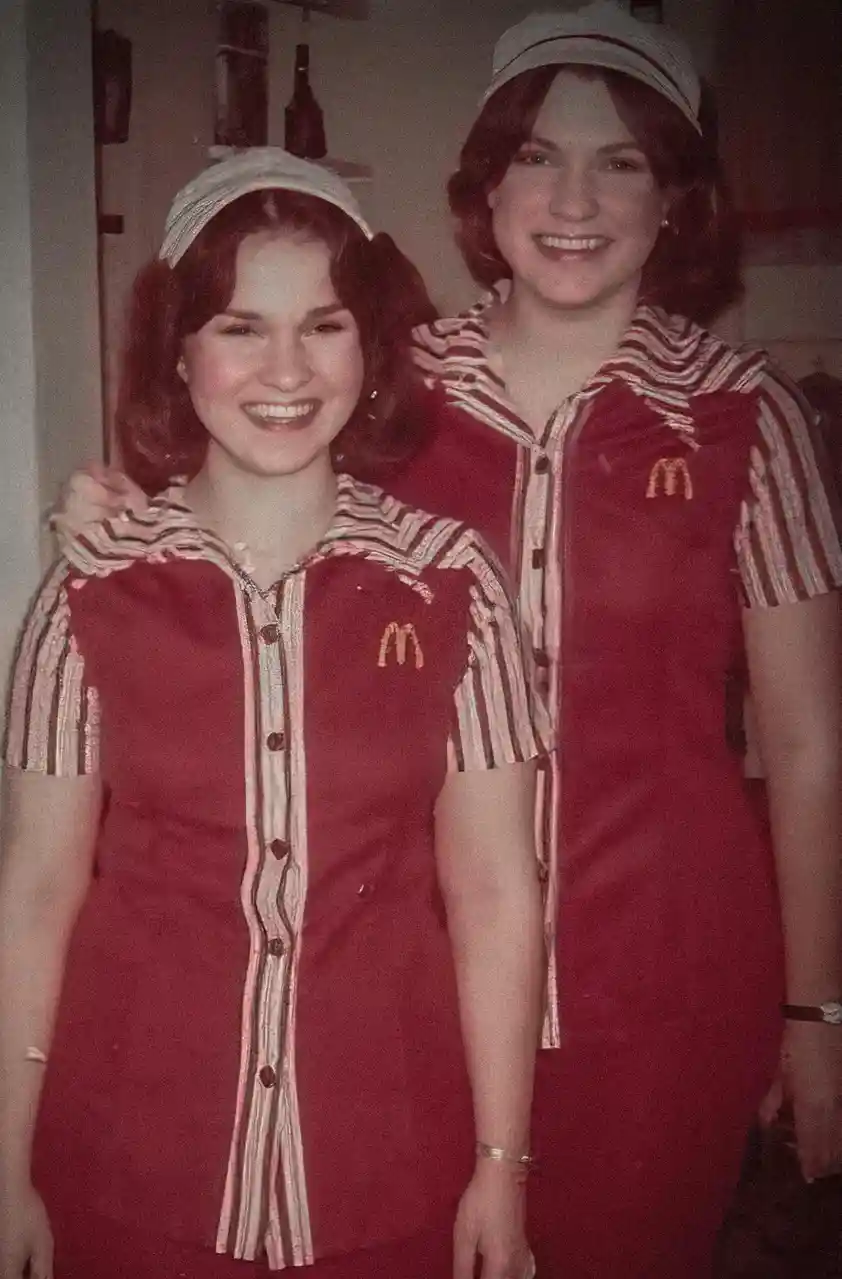 Two smiling women in vintage McDonald's uniforms with red dresses, striped collars, and matching hats pose for a photo. The…