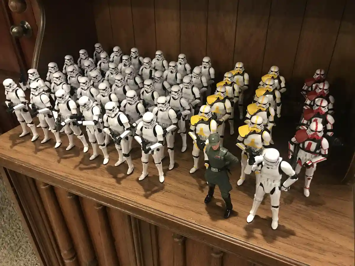 A collection of Star Wars action figures, mostly white-armored Stormtroopers, are arranged on a wooden shelf. A single…