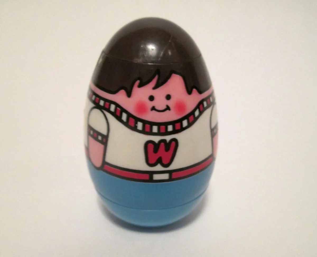 A close-up of a plastic egg-shaped toy with a cartoon character painted on it, featuring a smiling face with black hair…