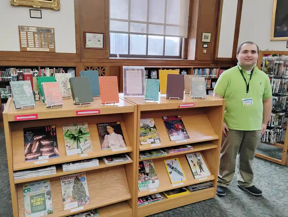 A man named Jacob, wearing a green polo shirt and a library lanyard, stands beside a display of vintage books and magazines…