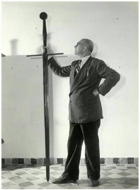 A black-and-white photo shows a bald man in a suit and tie standing next to an enormous sword mounted vertically on a wall…