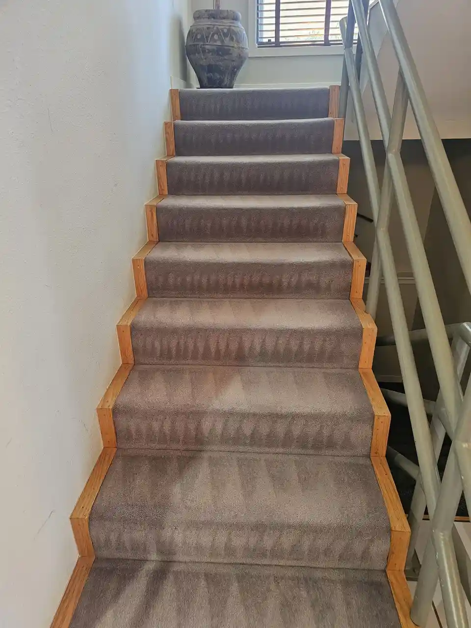 A carpeted staircase with wooden risers and a metal railing leads upward, ending at a large decorative vase near a window…