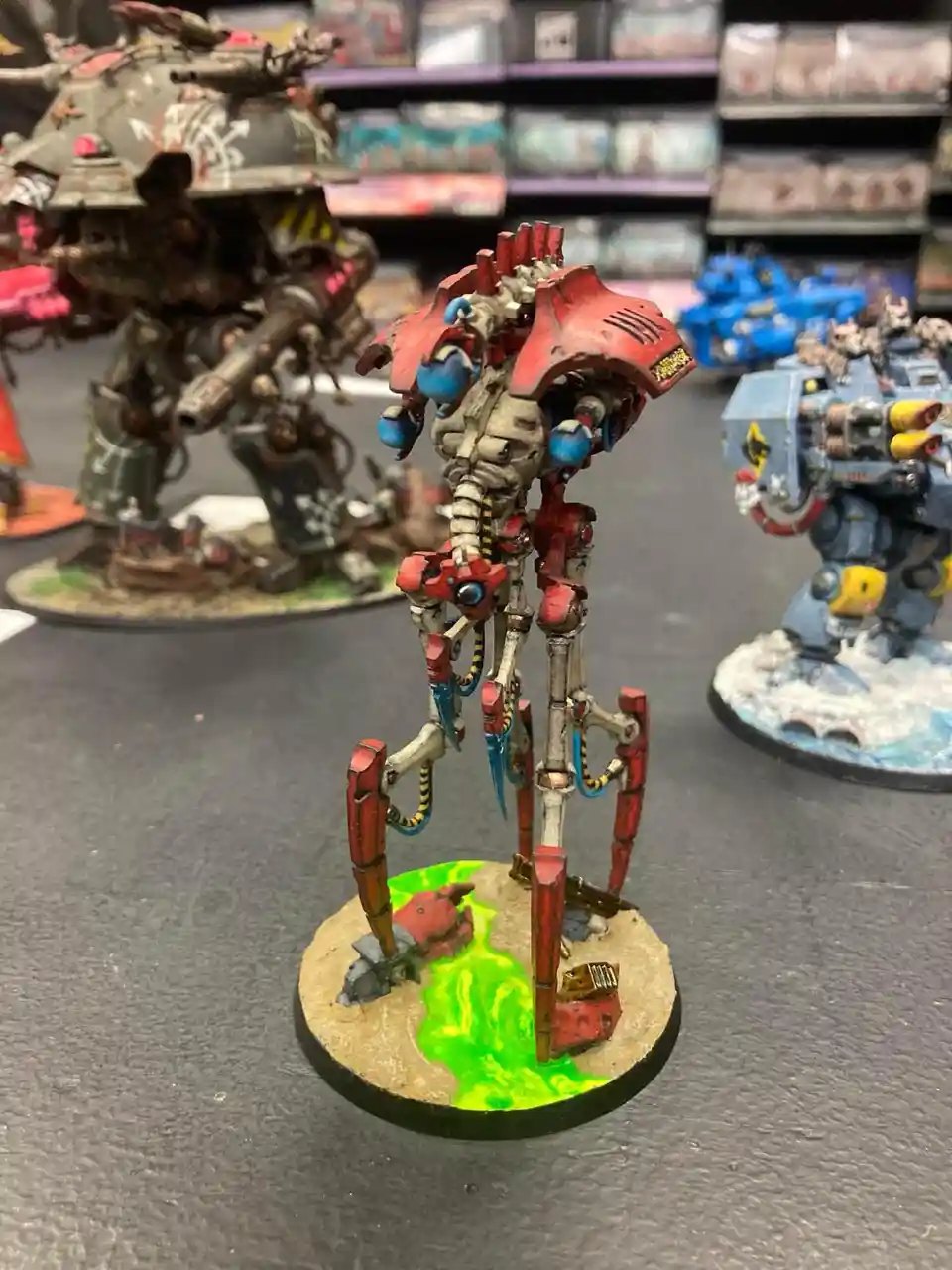 A close-up photo of a painted, red and grey biomechanical robot miniature standing on a circular base with green glowing…