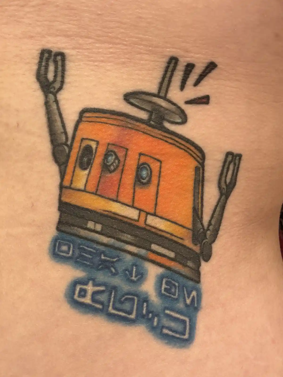 A tattoo on skin depicts a cartoonish orange cable car with fork-like arms and a spinning wheel on top, emitting sound…