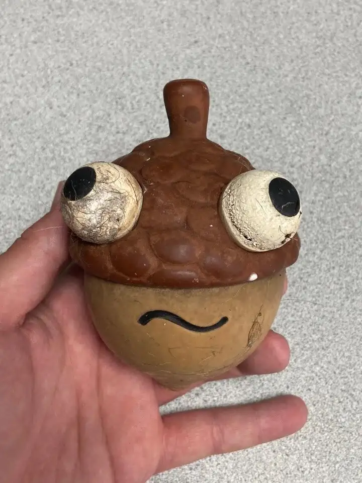 A hand holds a handmade clay acorn figurine with large, googly eyes and a wavy black smile drawn on its lower half. The…